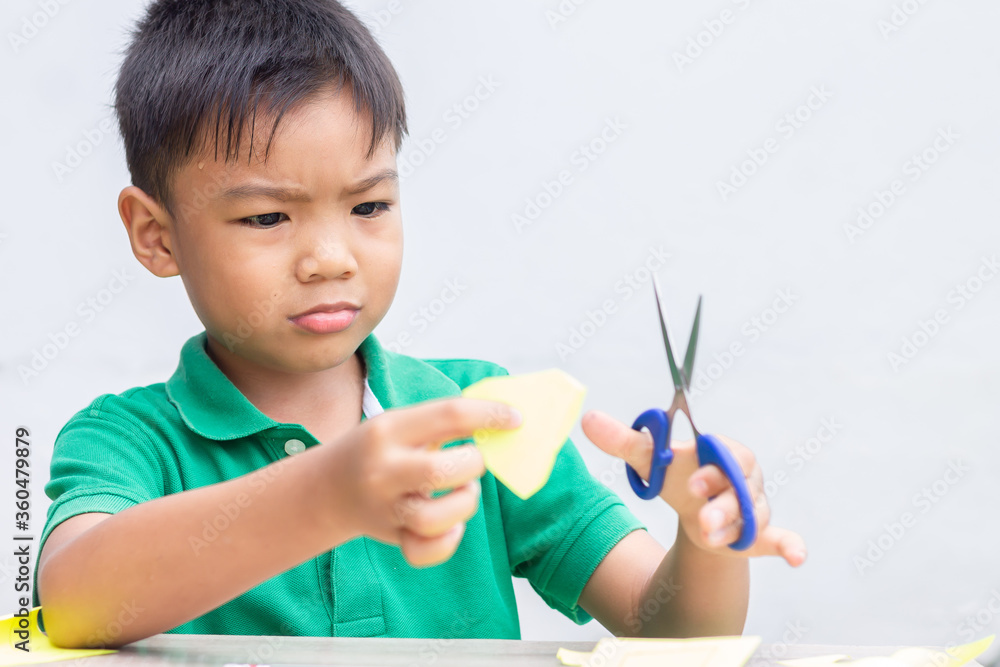 Selective focus. Portrait image of 5-6 years old Asian child boy practice to cutting color paper by the scissors on the wooden table.Study from home, social Distancing, Kid and education concept. Selective focus. Portrait image of 5-6 years old Asian child boy practice to cutting color paper by the scissors on the wooden table.Study from home, social Distancing, Kid and education concept.