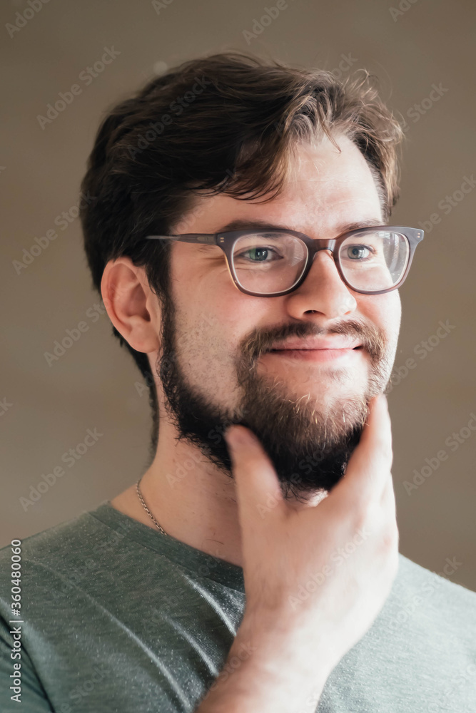 Obraz premium a handsome young man with a thick beard and glasses is illuminated on one side by a soft light. fashionable beard, satisfied