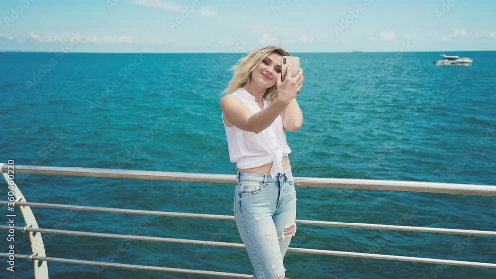 Young attractive blonde girl in a white blouse and blue jeans, makes selfie standing on the wooden pier on sunny summer day on the background of the sea. Young attractive blonde girl in a white blouse and blue jeans, makes selfie standing on the wooden pier on sunny summer day on the background of the sea.