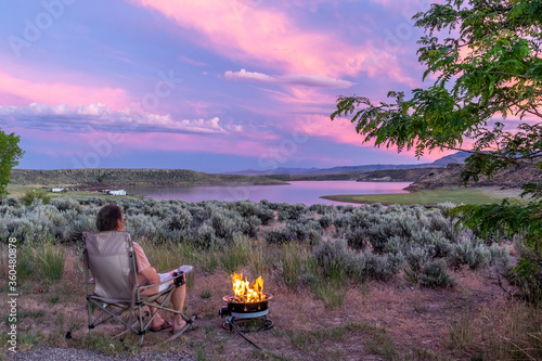 A man sitting by a burning fire watching a scene with sage brush, a tree, a lake and mountains while the Belt of Venus is painting the clouds pink and red as the sun is setting over the horizon, photo