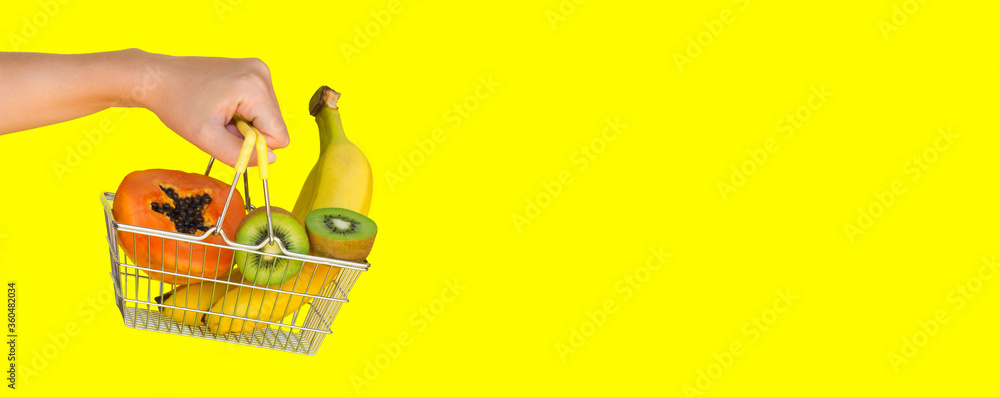 A female hand holding a shopping cart or basket filled to the top with fresh fruits (banana, kiwi, papaya) on a yellow background. The concept of buying food and fruit online. Diet food. Fast delivery