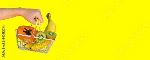 A female hand holding a shopping cart or basket filled to the top with fresh fruits (banana, kiwi, papaya) on a yellow background. The concept of buying food and fruit online. Diet food. Fast delivery