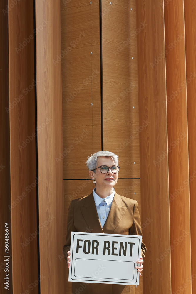 Portrait of mature businesswoman in eyeglasses holding placard For Rent while standing against the office building Portrait of mature businesswoman in eyeglasses holding placard For Rent while standing against the office building