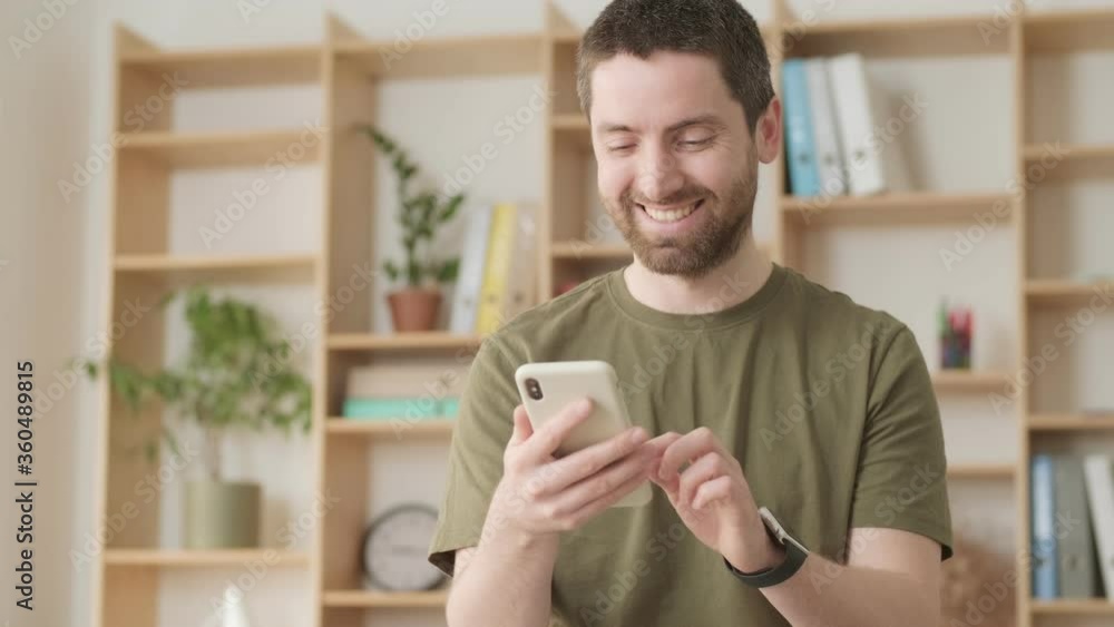 A smiling positive man is typing on his smartphone standing in the office A smiling positive man is typing on his smartphone standing in the office