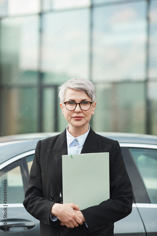 Fototapeta premium Portrait of mature businesswoman in suit and in eyeglasses holding documents and looking at camera while standing in the city