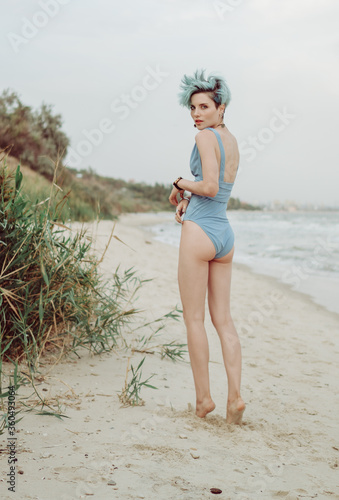 Young attractive girl with turquoise hair in a blue bathing suit posing on the seashore near the green thickets of reeds
