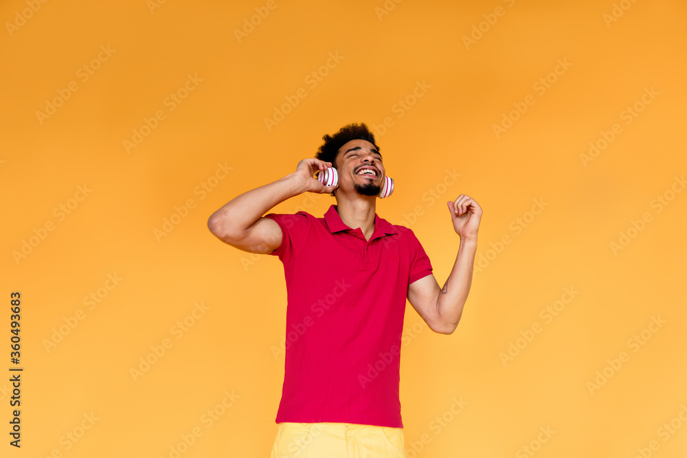 Indoor photo of happy exited man in headphones wearing red shirt and yellow trousers. Studio shot of chilling afro american guy. Indoor photo of happy exited man in headphones wearing red shirt and yellow trousers. Studio shot of chilling afro american guy.