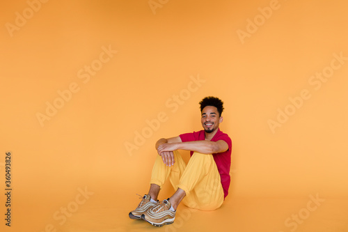 Full-lenght portrait in studio wearing red t-shirt and yellow trousers sitting over yellow background. Happy emotions, isolated background