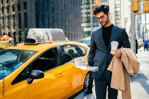 Handsome male in elegant suit walking on Manhattan near yellow cab reading information from daily press, prosperous businessman in trendy outfit checking financial article