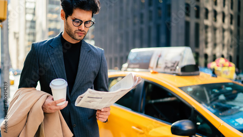 Puzzled proud CEO dressed in elegant wear reading business publication standing on Manhattan street, successful entrepreneur with coffee takeaway searching financial news in newspaper in downtown