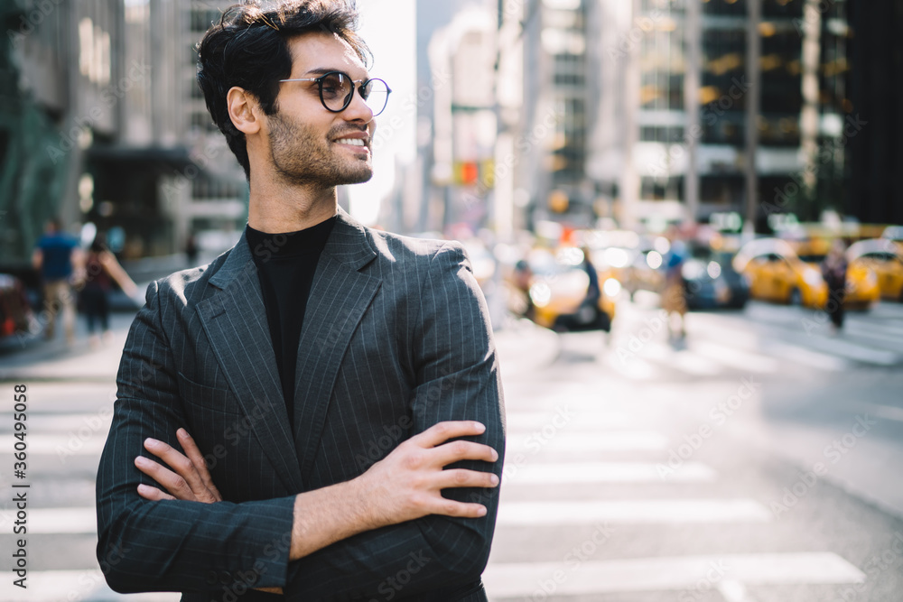 Side view of cheerful handsome man in stylish formal wear and spectacles looking away on Manhattan, prosperous proud ceo in elegant suit standing with crossed arms smiling on urban setting background.