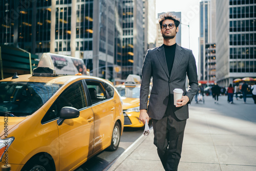 Serious male manager 30s dressed in elegant trendy wear and eyeglasses spending time outdoors, handsome businessman with coffee to go walking near traffic road with taxi in metropolitan downtown © BullRun