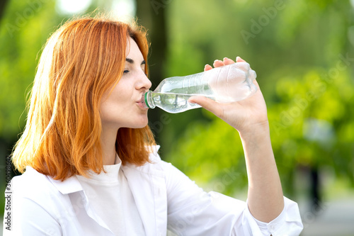 Young redhaired woman drinking water from a bottle on summer day.