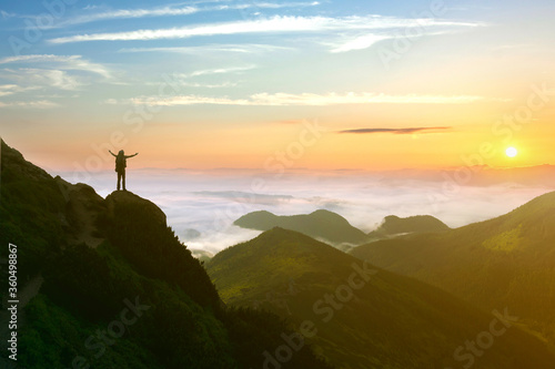 Small silhouette of hiker man enjoying beautiful sunrise in morning mountains. Traveler with raised hands standing on mountain with white fog below. © bilanol