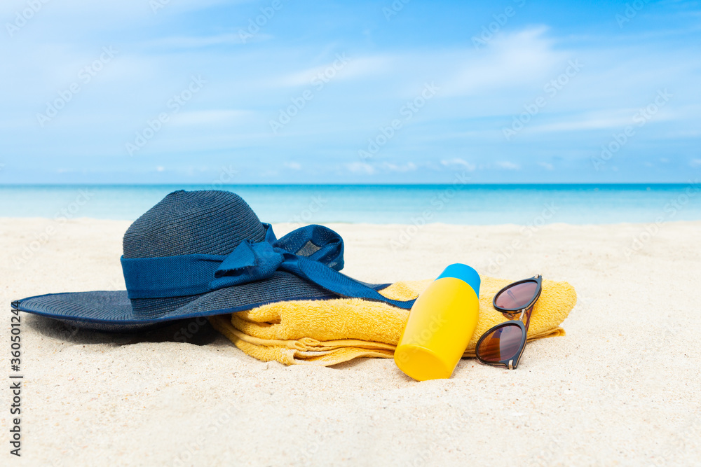 Dark blue hat, sunblock lotion bottle and sunglasses lie on yellow towel on tropical white sand beach with sea shore background. Vacation concept in summer time.