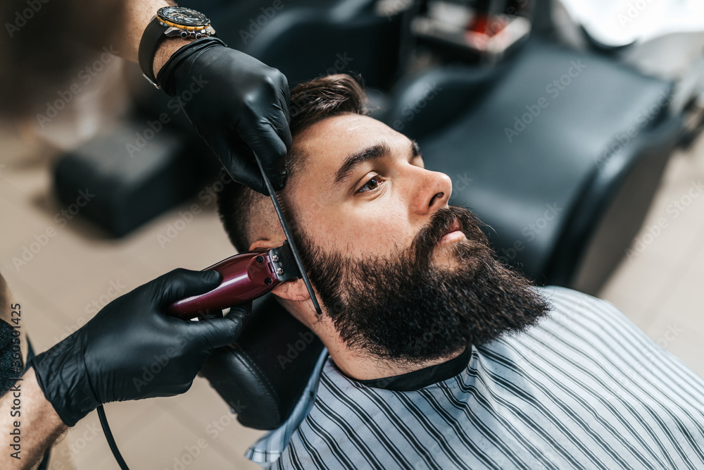 Young good looking man visiting barber shop. Trendy and stylish beard styling and cut. - obrazy, fototapety, plakaty 