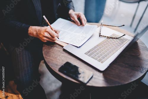 Cropped image of man making financial calculations in business planning of banking company sitting indoors, stylish male lawyer working with documents checking legal information in cafe interior photo