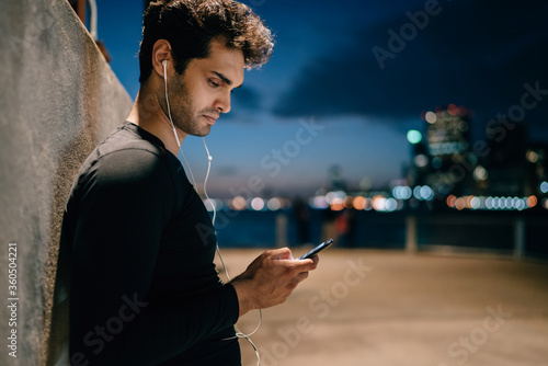 Side view of fit man dressed in tracksuit listening music in modern earphones connected to smartphone standing on evening city promotional background after workout in urban setting