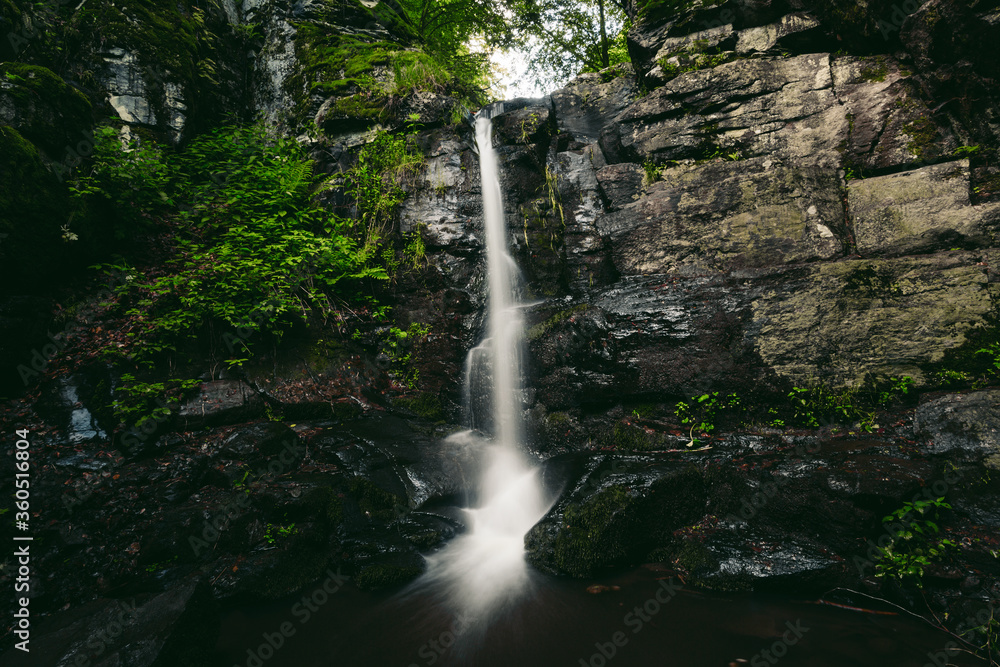 Naklejka premium Beautiful view of moody and dramatic waterfall with big rocks and stones on foreground in forest. Small dark waterfall in European forest - silky smooth effect. Waterfall in the jungle - green leafs.