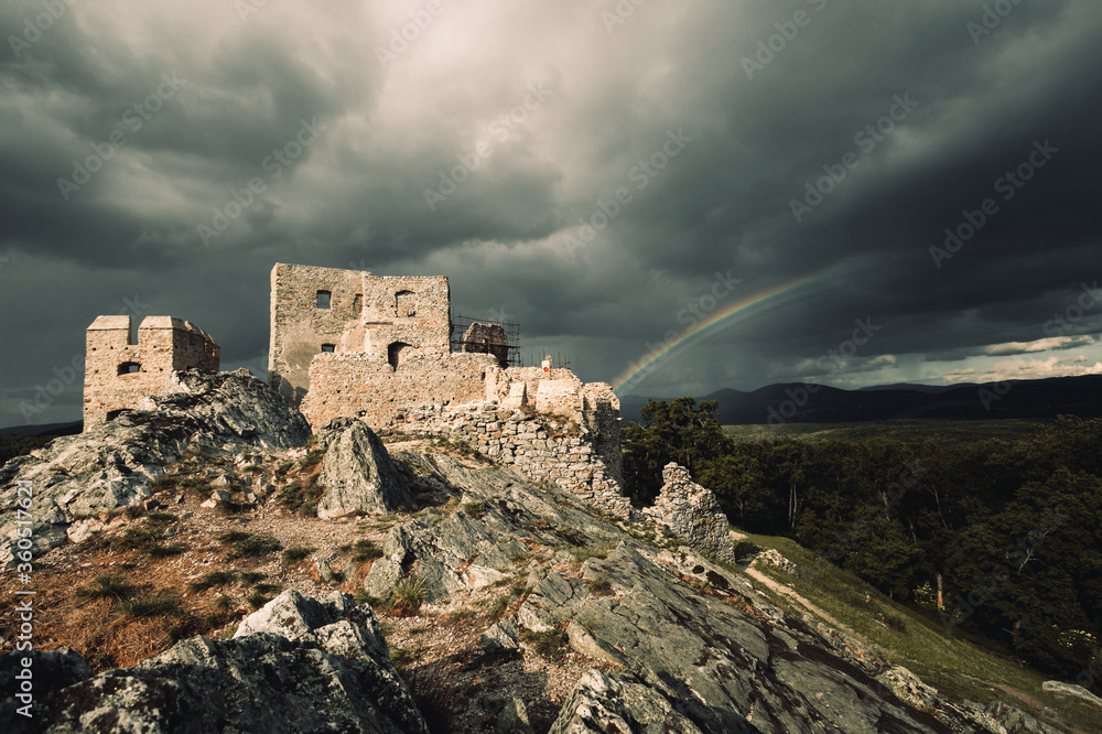 Dark and moody view of Hrusov Castle in Europe (Slovakia) before the storm. Old Ruins of castle on the dark rocks with rainbow on background.  Dramatic shot of castle on the hill with misty sky.