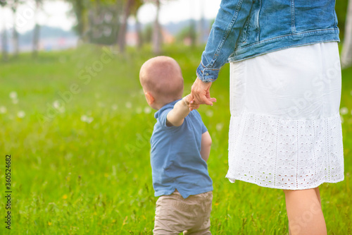 Close up of a woman holding a child by the hand near her, view from the back. The kid walks with mom on the green grass.