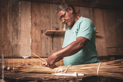 Traditional handicraft mat made of dry reed in a local community of Garopaba, Brazil. Artisan working at his wooden workshop. Craftsman wearing green t-shirt photo