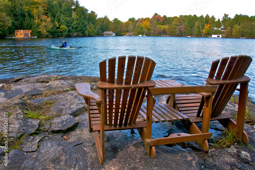 Retirement Living - Two Muskoka chairs sitting on a rocky shore facing a calm lake with cottages in the background