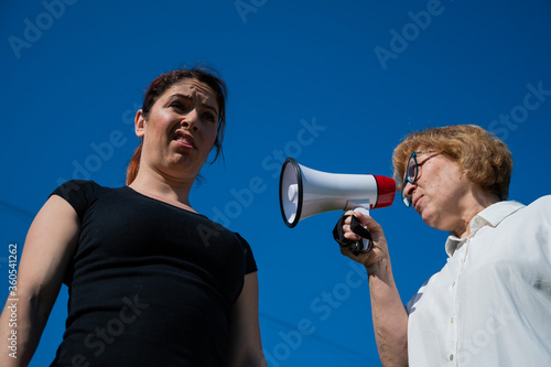 The conflict of generations. An emotional elderly woman shouting at her daughter in a megaphone. An elderly mother swears at a middle-aged woman on a loudspeaker on a blue background. © Михаил Решетников