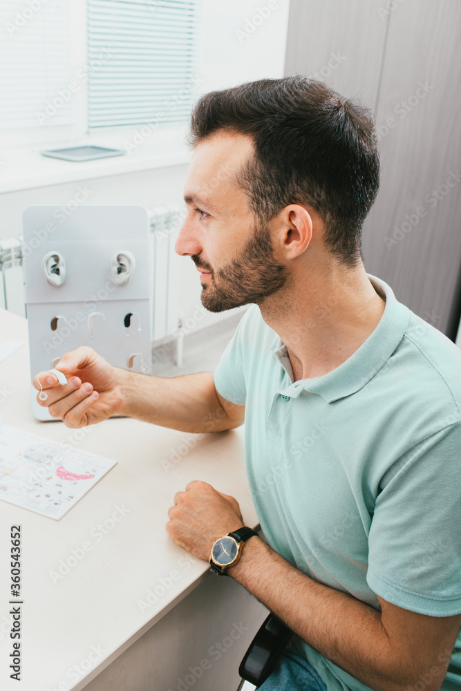 Close-up portrait of an adult male with hearing impairment during the selection of hearing aids in a hearing clinic. Hearing impairment treatment, hearing solutions Close-up portrait of an adult male with hearing impairment during the selection of hearing aids in a hearing clinic. Hearing impairment treatment, hearing solutions