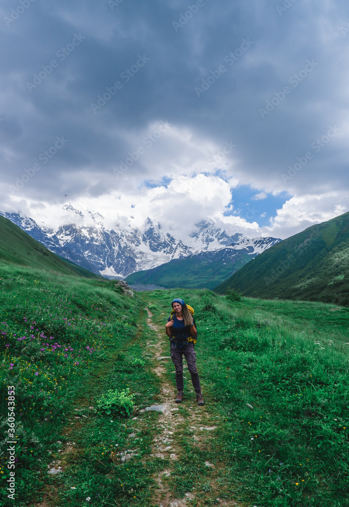 Girl traveler with a backpack in the mountains of Georgia. North Caucasus. Walking route to the village of Ushguli. Green valley with wild flowers.