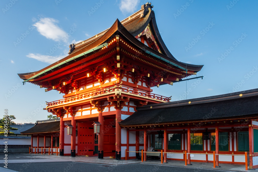 View of red entry gate to Fushimi Inari-Taisha Shinto Shrine in Kyoto, Japan, illuminated by morning sun. 