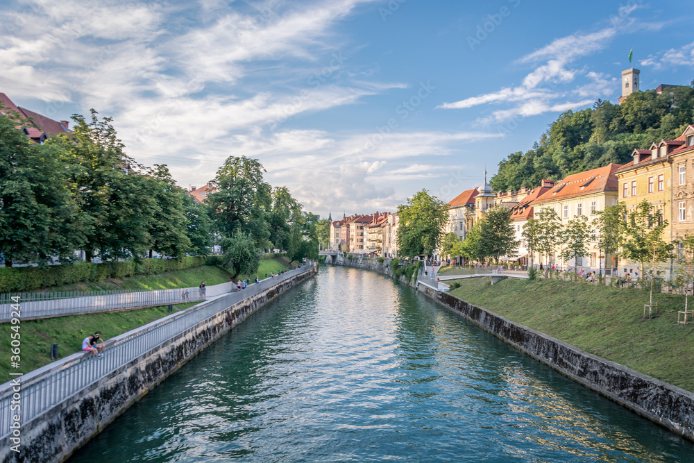 Elevated view of Ljubljanica river on sunny day. Summertime in Ljubljana city, Slovenia, full of tourists. River boat transports tourists. Historic buildings and castle in background