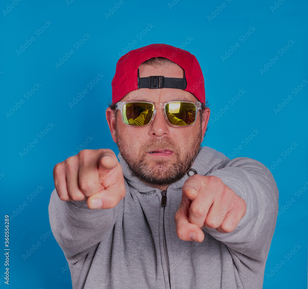 Guy with beard ,cap and sunglasses on pointing ,shot on a blue background. Guy with beard ,cap and sunglasses on pointing ,shot on a blue background.