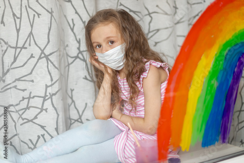 A child in a medical mask looks out the window through a painted rainbow on glass. A flash mob, a child draws a rainbow on a window pane during world quarantine due to coronavirus infection..