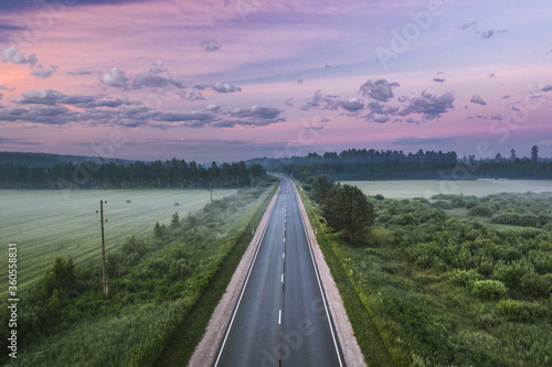 Highway going through meadow in mist surrounded by forest. Aerial view over picturesque landscape in fog at sunrise. 