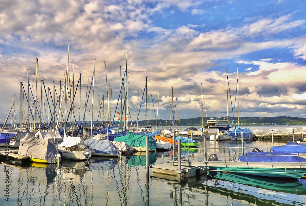 Fototapeta premium Sailboat harbor on Lake Constance, in the background you can see the Lake Constance ferry. The sky has many white clouds on blue, in the water you can see the mirrored boats.