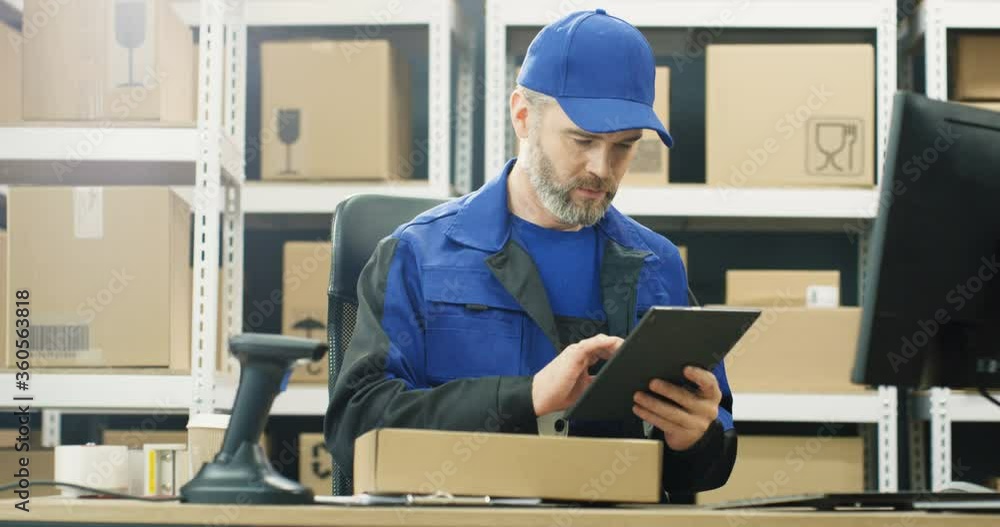 Handsome Caucasian mailman in uniform and cap sitting at table in postal office store and working at tablet device. Male courier filling in invoice and entering data while tapping on computer screen. Handsome Caucasian mailman in uniform and cap sitting at table in postal office store and working at tablet device. Male courier filling in invoice and entering data while tapping on computer screen.