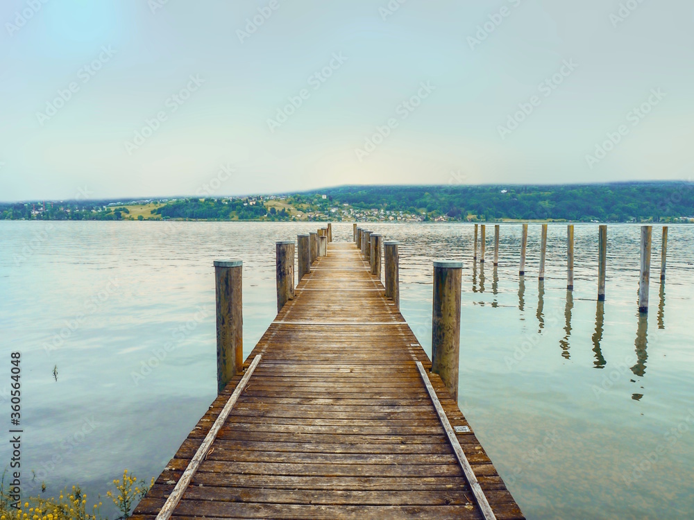 A jetty of wooden planks protrudes into Lake Constance (Germany). The almost light turquoise evening sky is reflected in the water, on the opposite side you can see a strip of land.