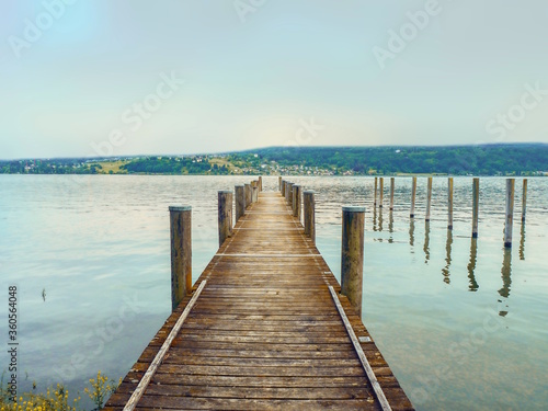 A jetty of wooden planks protrudes into Lake Constance (Germany). The almost light turquoise evening sky is reflected in the water, on the opposite side you can see a strip of land. © AkimaFutura