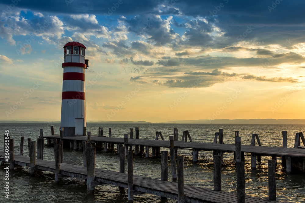 A wooden pier leading to the lake and a red and white lighthouse on Lake Neusiedl in Podersdorf, Austria. In the background is a dramatic sky at sunrise. A wooden pier leading to the lake and a red and white lighthouse on Lake Neusiedl in Podersdorf, Austria. In the background is a dramatic sky at sunrise.