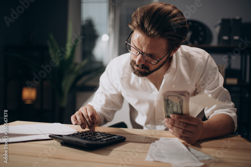 Serious businessman in formal clothing and eyeglasses using calculator for counting money and comparing with all bills that lying on table. Concept of finance and savings.