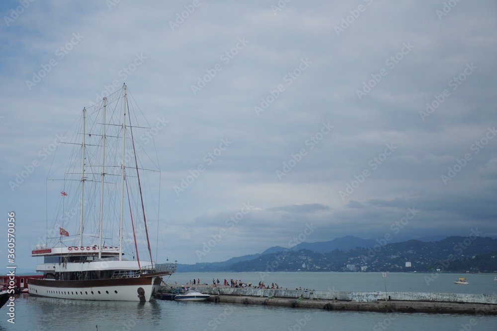 Sailboat with Turkish and Georgian flags and lowered sails at the pier against the backdrop of mountains and a thunderstorm sky