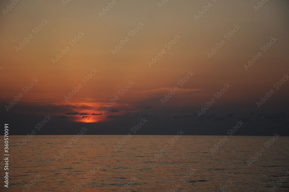 The rays of the setting sun behind small dark clouds over a calm sea