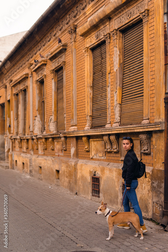 A beautiful caucasian girl with sunglasses stands next to her dog breed American Staffordshire Terrier in front of an old house on the street
