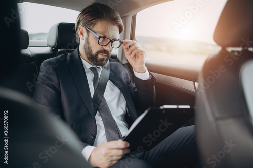 Serious bearded man in formal clothing reading documents while sitting on back seat at luxury car. Mature businessman in eyeglasses looking through contract on the way to meeting. © Kuz Production