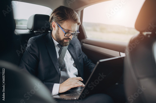 Handsome businessman in trendy suit sitting on backseat in car and working on wireless laptop. Busy bearded man in eyeglasses solving business issues online.