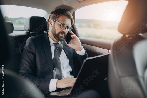 Concentrated businessman in eyewear and black suit having conversation on mobile phone while riding in car. Mature man sitting on backseat with opened laptop on knees. © Kuz Production