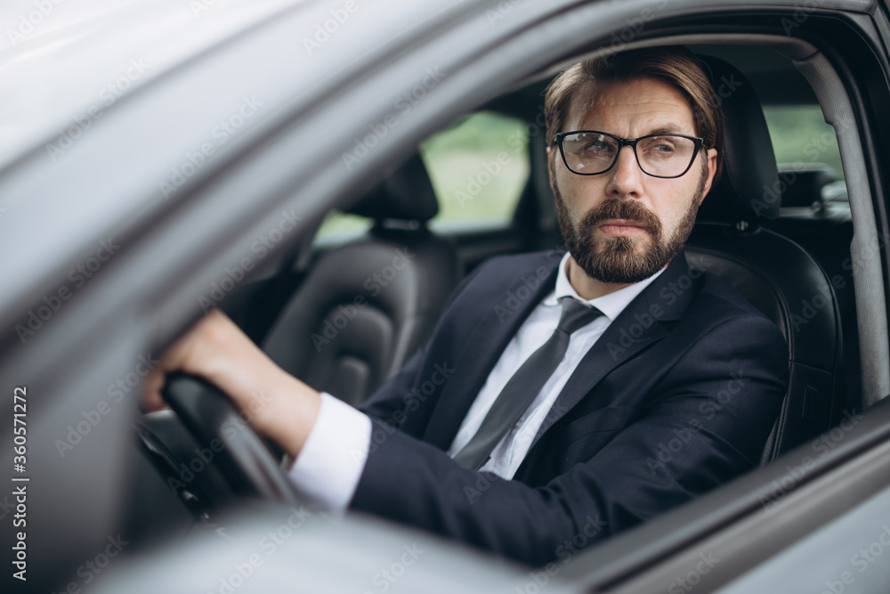 Portrait of confident bearded man wearing eyeglasses and black suit driving own luxury car. Successful businessman is hurry to important meeting. Portrait of confident bearded man wearing eyeglasses and black suit driving own luxury car. Successful businessman is hurry to important meeting.