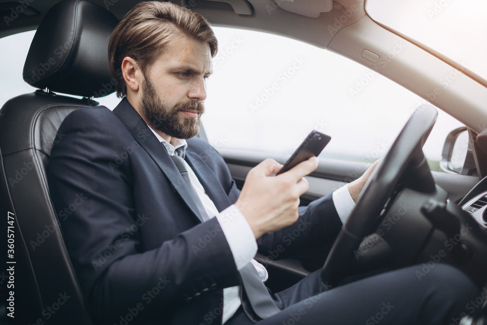 Side view of busy bearded man dressed in formal clothing using smartphone while driving car. Mature businessman texting message while sitting in automobile.