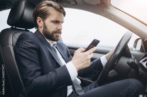Side view of busy bearded man dressed in formal clothing using smartphone while driving car. Mature businessman texting message while sitting in automobile. © Kuz Production